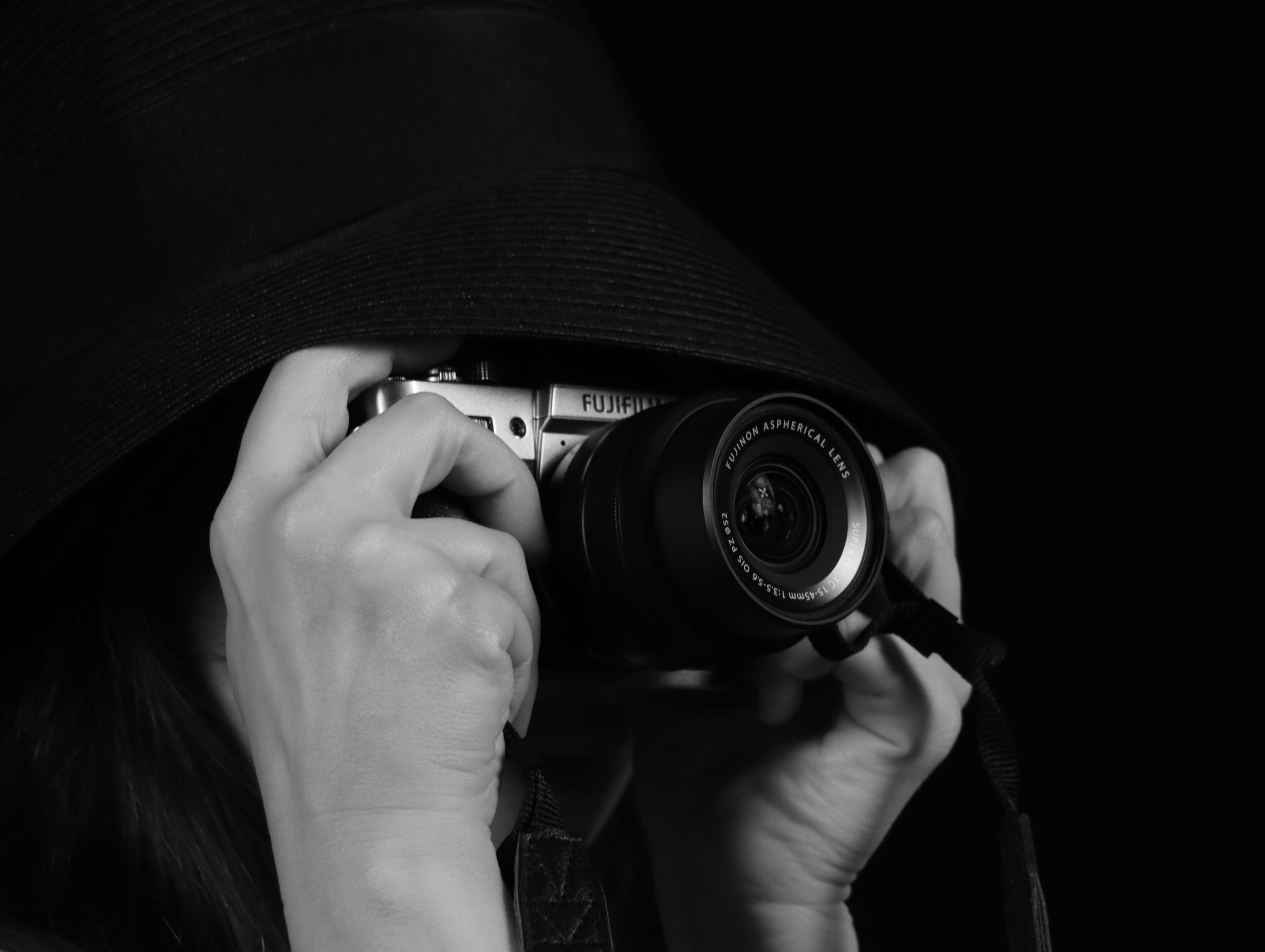 Black and white portrait of a person using a camera partially hidden by a hat.